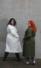 Fototapeta na wymiar Two plus-size girls in coats standing against a gray wall.