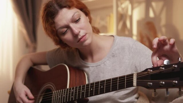 Music hobby. Guitar adjusting. Instrument entertainment. Concentrated woman student guitarist tuning pegs and gears on headstock at home.