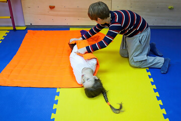 boy and girl children roll each other up in a thick blanket on mats controlling a small space for...