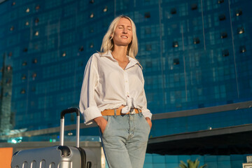 Portrait of a young happy blonde woman in a white shirt and jeans with a suitcase with her eyes closed against the background of a modern glass hotel in the rays of the sun, copy space