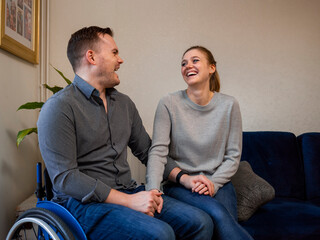 Woman and man on wheelchair holding hands and laughing at home