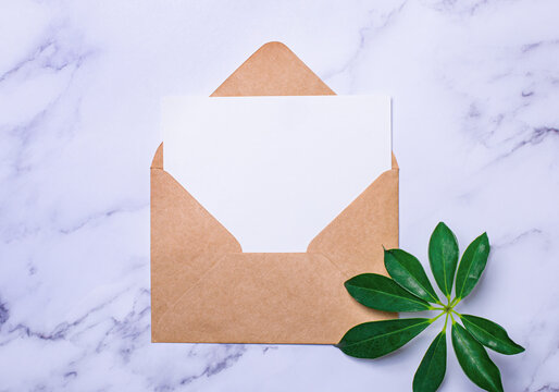 On a light marble background there is a craft envelope, a sheet of sheflera and a white sheet of paper with a place to insert text. Copy space