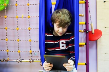 child boy watching cartoons on a tablet while sitting in a hammock at a children's center for the development of special children