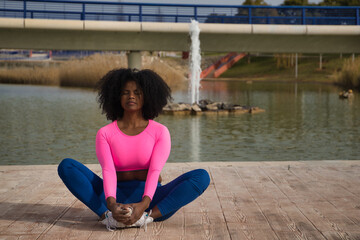 African-American woman with afro hair and sportswear, with fluorescent pink t-shirt and leggings, doing yoga and relaxation exercises by an outdoor lake. Fitness concept, sport, street, urban.