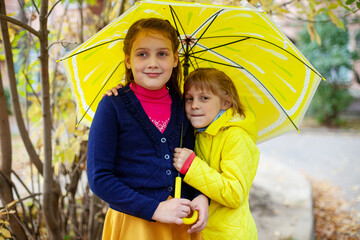  Two girls of 7 and 10 years old   hugging under   yellow umbrella in autumn.