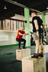 Three sportsmen performing jumping squats on wooden cubes in contemporary crossfit gym. Focus on athlete in red t-shirt and black leggings doing exercise with concentration.