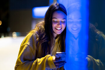  Friendly young woman with long hair next to a bluish neon wall looking at her mobile phone dressed...
