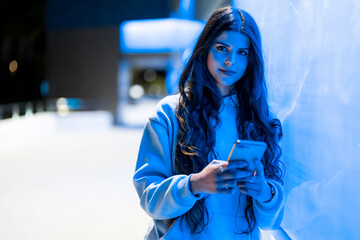 Attractive and friendly young woman with long hair next to a bluish neon wall looking at her mobile...