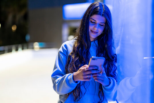Attractive and friendly young woman with long hair next to a bluish neon wall looking at her mobile phone dressed in a hoodie