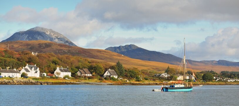 Traditional English gaff cutter with a motorboat sailing near the shore of a small town Craighouse. Mountains peaks of Paps of Jura in the background. Dramatic sky. Inner Hebrides, Scotland, UK