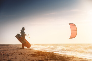 Kiteboarding. Kitesurfing athlete woman at sunset stands on the sandy shore holding her kite in the air and looks at the sea with waves and sunset - obrazy, fototapety, plakaty
