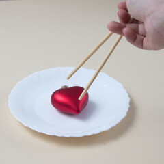 A heart on a white plate and Chinese sticks held by a hand on right side trying to catch it. Minimal love scene.