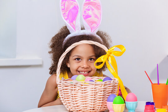 Little Black girl with bunny ears hiding behind Basket full of colored eggs
