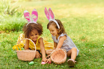 two girls are sitting on the green lawn during easter egg hunt and putting Easter eggs in baskets