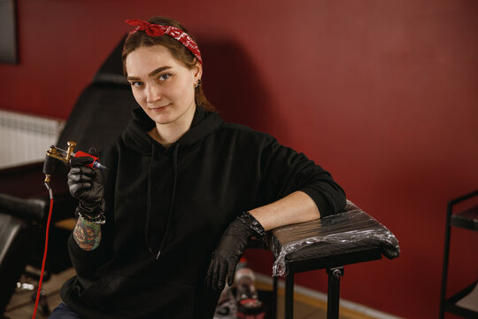 Portrait of young lady tattoo master wearing black hoodie and gloves sitting in tattoo salon with tattoo machine in hands