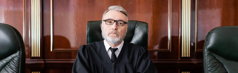 mature and serious judge in eyeglasses looking at camera in court, banner
