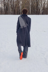 A young man in a long gray coat and scarf walks through the snow. Rear view. Back view.