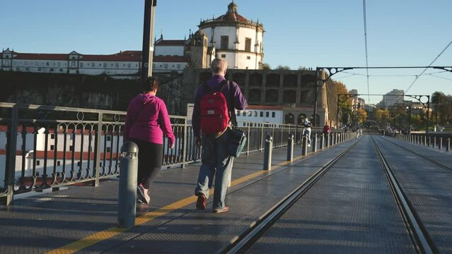 Elderly gray haired man with backpack walking on Ponte de Dom Luis I. European tourism place in Portugal, city of Porto. Old traveling tourists family couple. Visiting Portuguese cityscape landmarks.