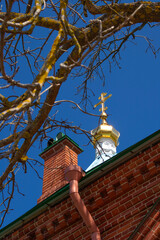 An Orthodox cross shining in the sun on the dome of the church. The dome is a brick building. In the foreground are blurred dry branches of a tree without leaves. Bottom view. Daylight. Spring.