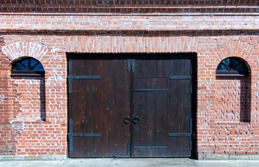 A closed double-leaf gate with a door in an old red brick building. Bricked-up windows. The gate is made of dark wood planks. Rings instead of handles on the gate. Summer. Hard daylight.