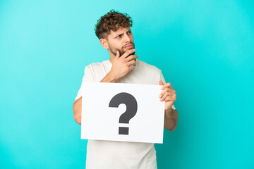 Young handsome caucasian man isolated on blue background holding a placard with question mark symbol and thinking