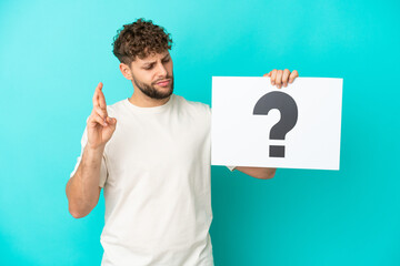 Young handsome caucasian man isolated on blue background holding a placard with question mark symbol with fingers crossing