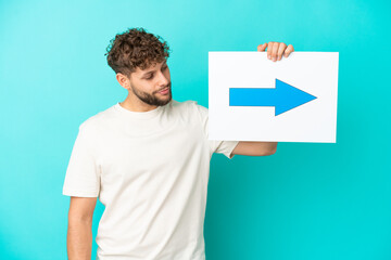 Young handsome caucasian man isolated on blue background holding a placard with arrow symbol