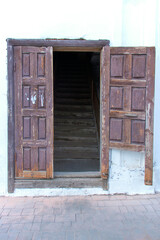 The entrance to an old dilapidated entrance with a broken wooden door. White walls. A wooden staircase is visible. Provincial building. Summer. Daylight.