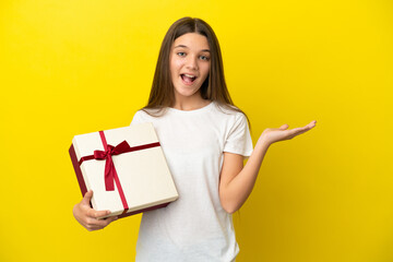 Little girl holding a gift over isolated yellow background with shocked facial expression