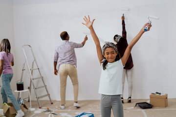 Little sweet adorable girl is happy to move to new apartment, helps in renovation of house, raises hands in the air with happiness, in the background parents and sister are painting walls of the room