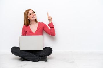 Young redhead woman with a laptop sitting on the floor isolated on white background showing and lifting a finger in sign of the best