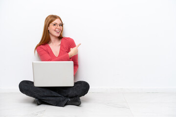 Young redhead woman with a laptop sitting on the floor isolated on white background pointing back