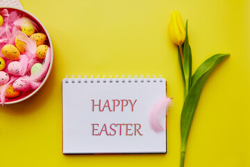 Happy Easter wishing. Pink bowl with colorful eggs and pink feathers. Post card on bright yellow background