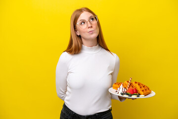 Young redhead woman holding waffles isolated on yellow background and looking up