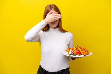 Young redhead woman holding waffles isolated on yellow background covering eyes by hands. Do not want to see something