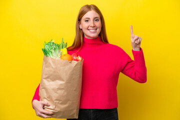 Young redhead woman holding a grocery shopping bag isolated on yellow background pointing up a great idea