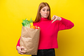Young redhead woman holding a grocery shopping bag isolated on yellow background showing thumb down with negative expression