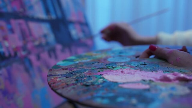 Focus on palette with oil paints. Artist draws picture on canvas in creative workshop. Woman dips brush into bright acrylic paints on palette and applies strokes to painting. Slow motion. Close up.