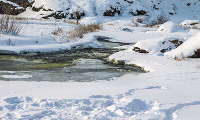 Winter landscape with a fast river with ice-free water, snow, dry grass and rocky shores