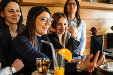 Group of adult caucasian women holding mobile phone while sitting at cafe or restaurant female millennial friends making a video call on smartphone or taking selfie photo real people leisure concept