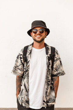 Happy young hipster man with smile in fashion summer clothes with bucket hat, shirt and sunglasses with backpack stands near white wall on the street