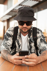 Cool hipster man in fashionable summer clothes with sunglasses sits outside and typing in his phone.