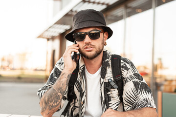 Handsome young stylish hipster guy in fashionable clothes with hats and sunglasses sitting on the...