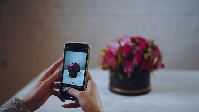 A girl takes pictures on her phone of a bouquet of flowers, a hatbox with flowers