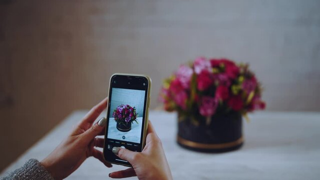 A girl takes pictures on her phone of a bouquet of flowers, a hatbox with flowers