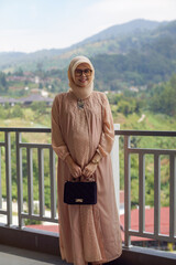 Portrait of pregnant young Asian muslim woman holding black hand bag standing by railing with mountain and blue sky in bokeh background. Smiling and happy expression.