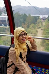 Portrait of young muslim woman wearing hijab sitting inside cable car with forest and mountain in background at Lake Ashi in Hakone, Japan. Smiling and happy expression.