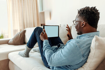 Shot of a handsome young man using his digital tablet while sitting on a sofa at home. Positive black man in casual reclining on sofa, using digital tablet, home interior.