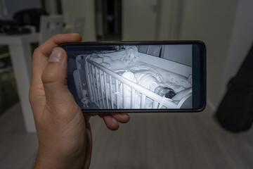 Unknown man holding mobile phone checking baby monitor watching his child sleeping via surveillance...