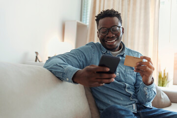 Focused smiling young man doing online banking with his phone while being seated on couch at home during the day. Young man using a smartphone and a credit card to shop online while sitting on sofa